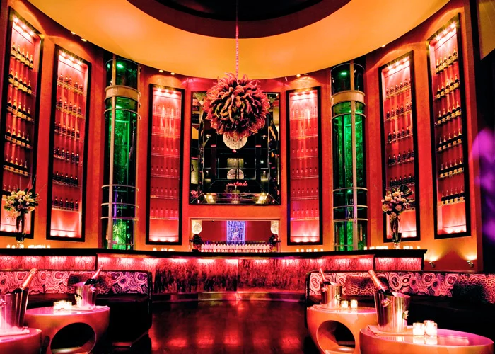 A vibrant bar interior with dramatic lighting, featuring colourful shelves lined with bottles, modern seating, and a striking central chandelier.